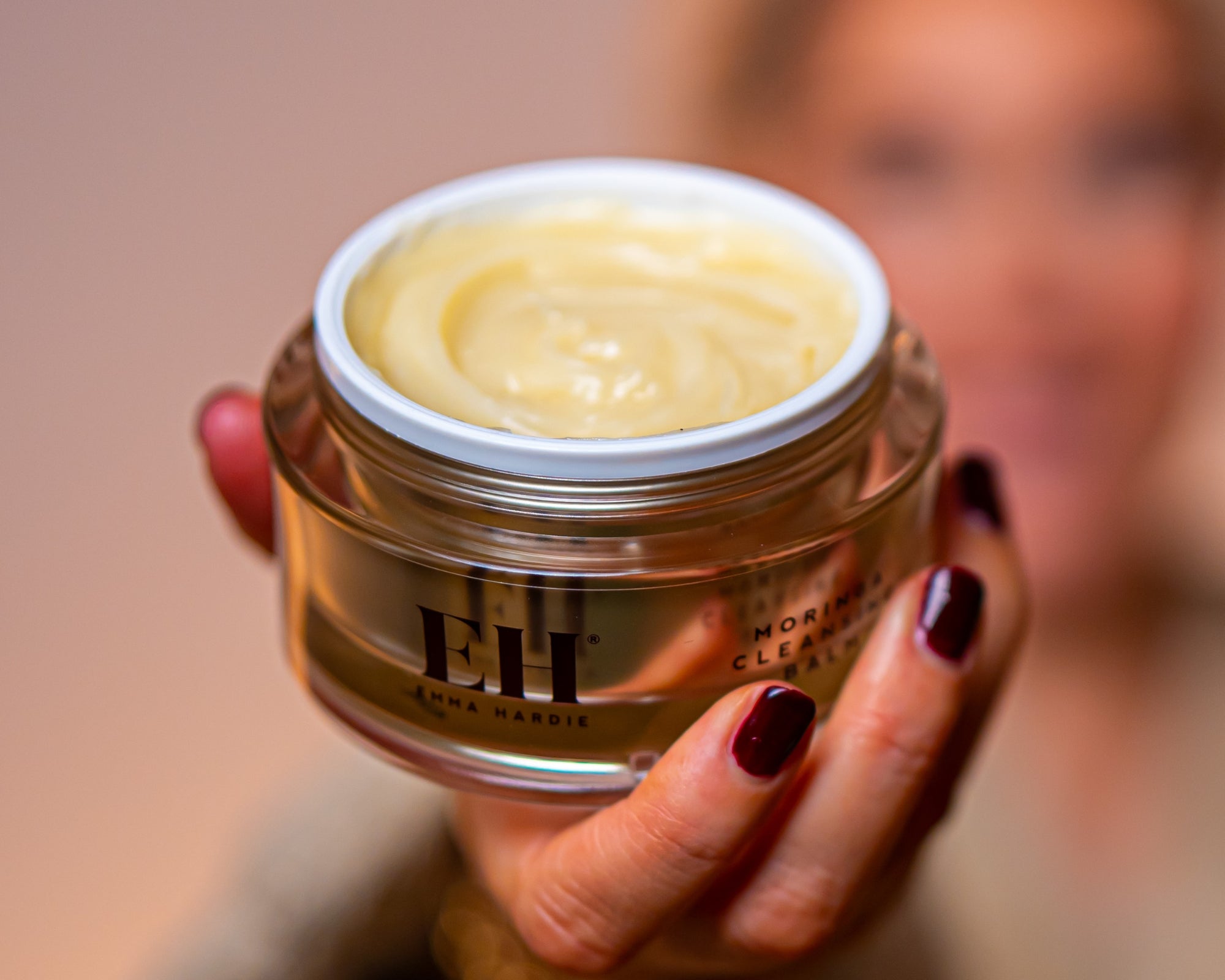 A close-up of a person holding an open jar of Emma Hardie Moringa Cleansing balm. The jar is clear glass showing the balm texture.