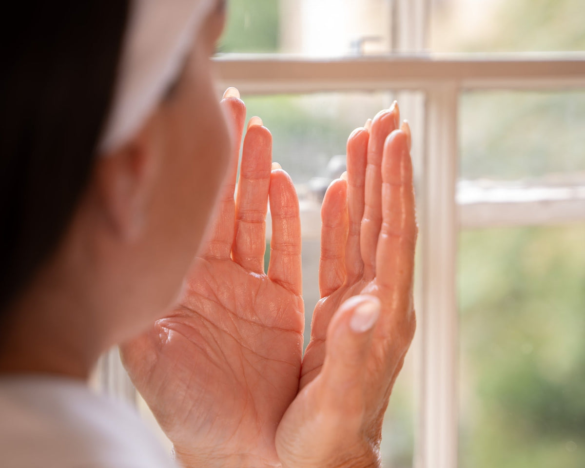 Hands gently pressing skincare into the face beside a sunlit window.