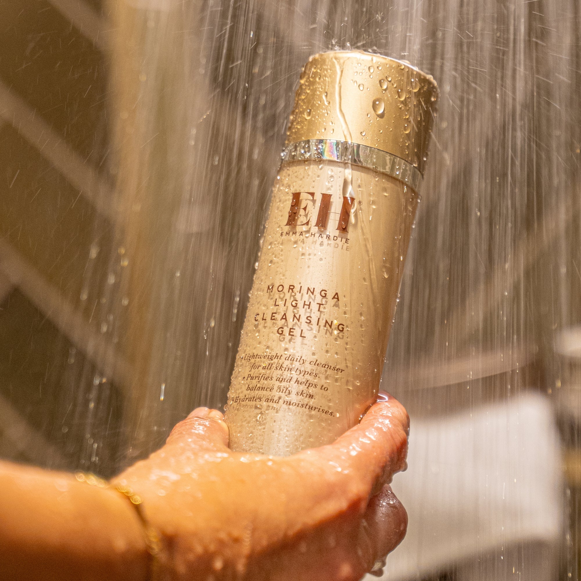 Hand holding Emma Hardie Moringa Light Cleansing Gel under running shower water, with water droplets across the bottle.