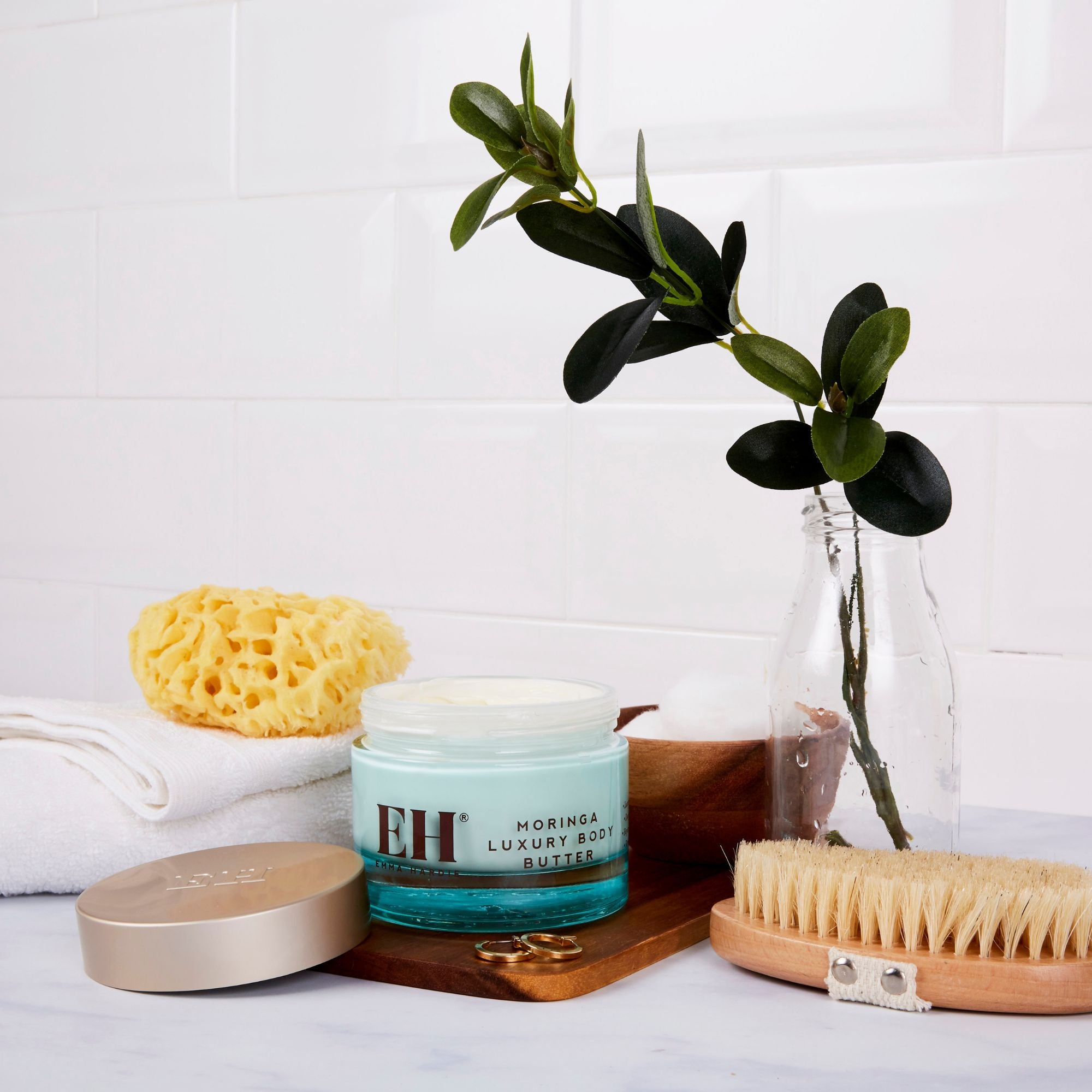 Emma Hardie Bath and body products including a jar of Moringa Luxury Body Butter, a sponge, and a brush on a white surface with a plant in the background.