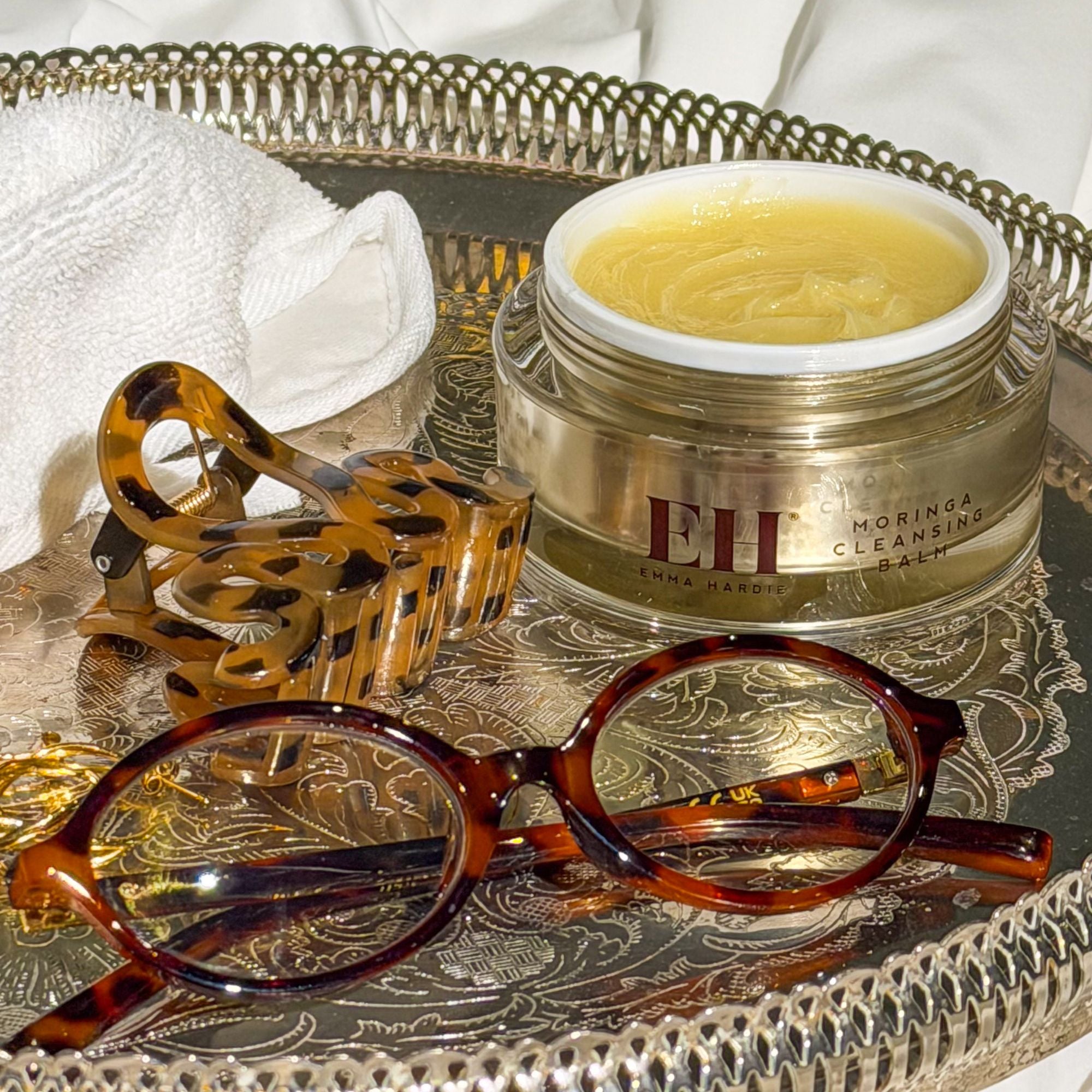 Open Jar of Emma Hardie cleansing balm with dual cleansing cloth on a silver tray with glasses and hair clip.