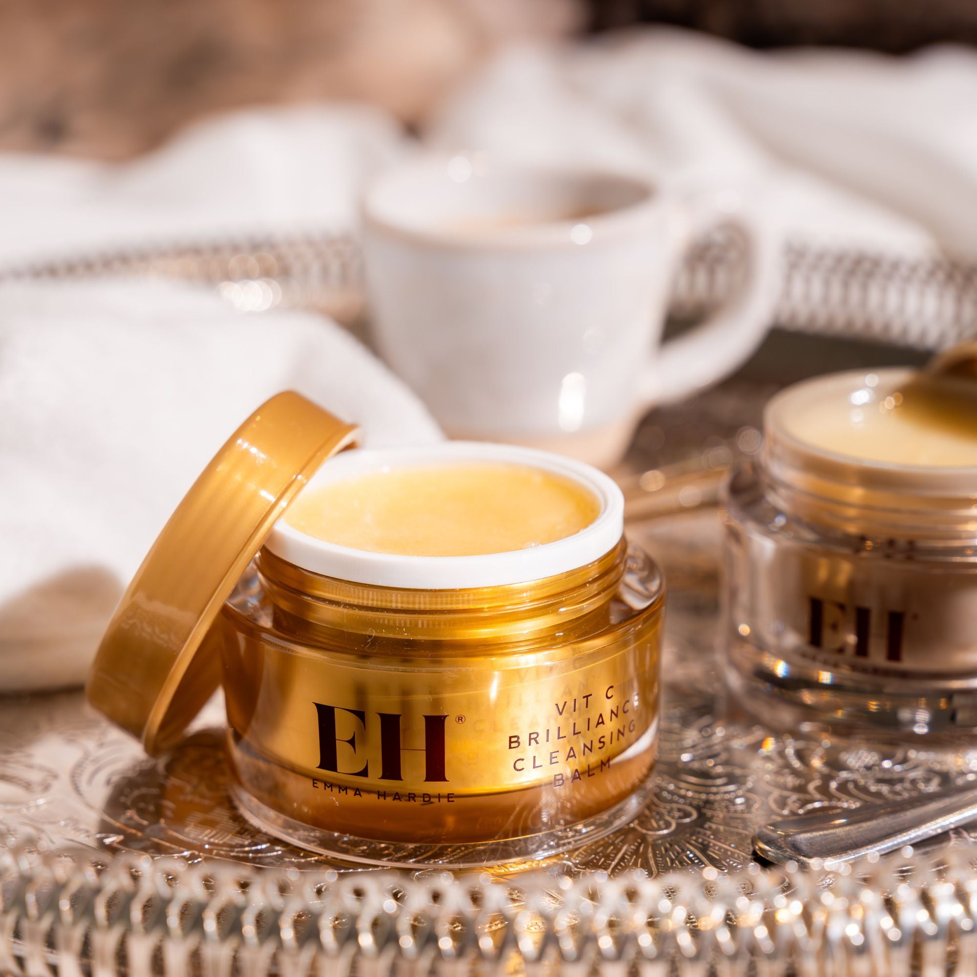 Emma Hardie Vit C Brilliance Cleansing Balm open on a decorative tray, with a soft towel, coffee cup and second balm jar in the background.