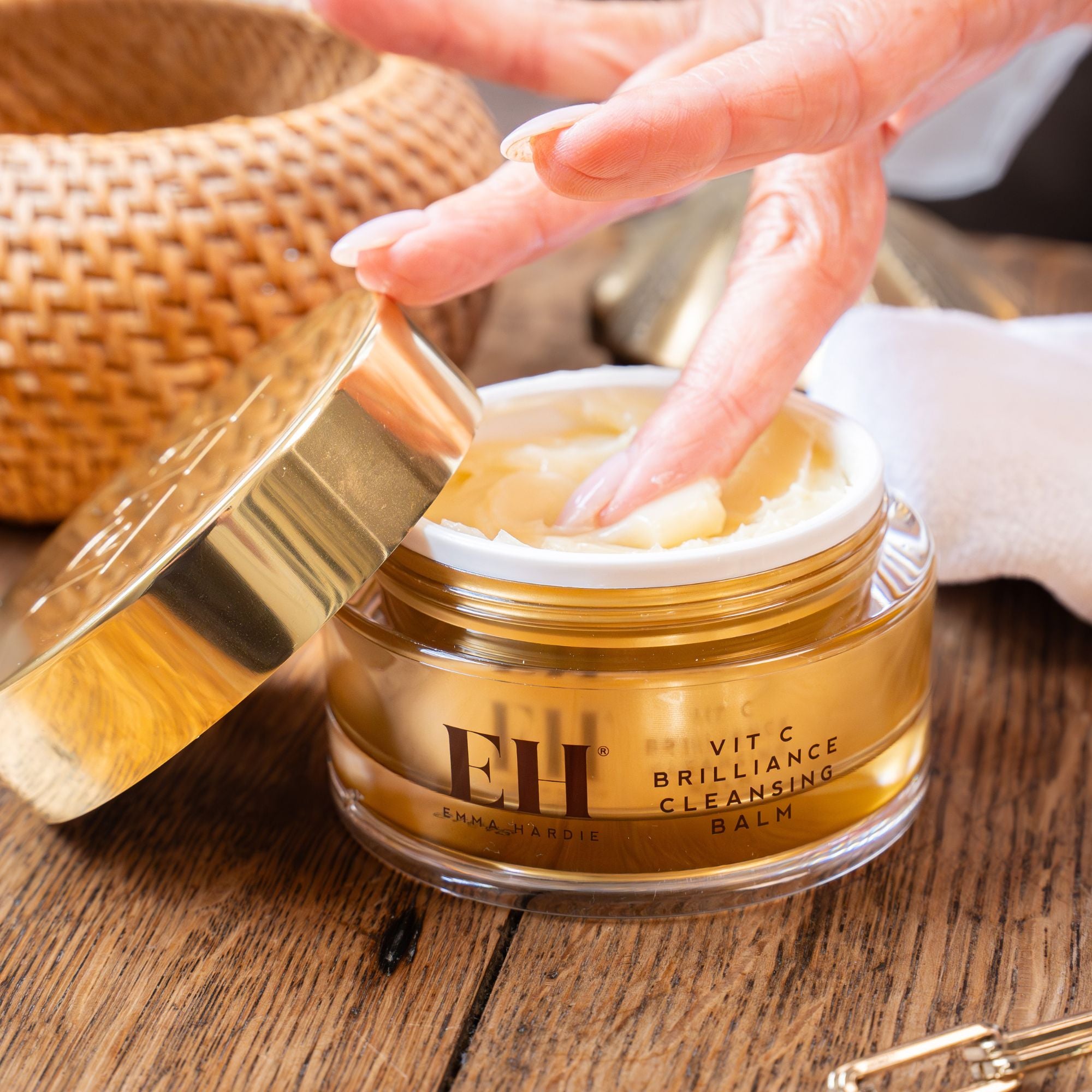 Fingertips scooping Emma Hardie Vit C Brilliance Cleansing Balm from an open gold jar on a wooden surface.