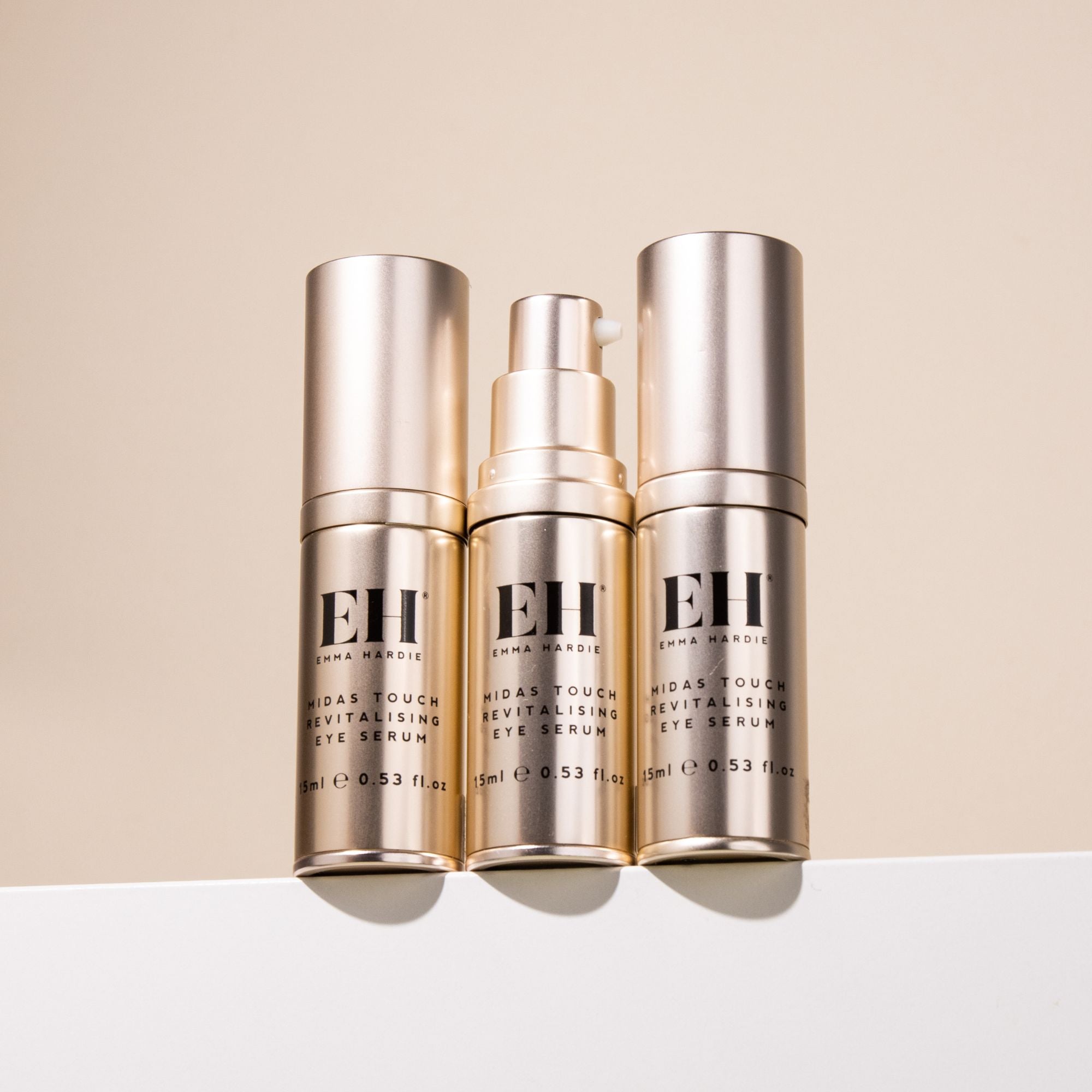 Three Emma Hardie Midas Touch Revitalising Eye Serum bottles against a soft neutral background, with one pump bottle open to show the product.