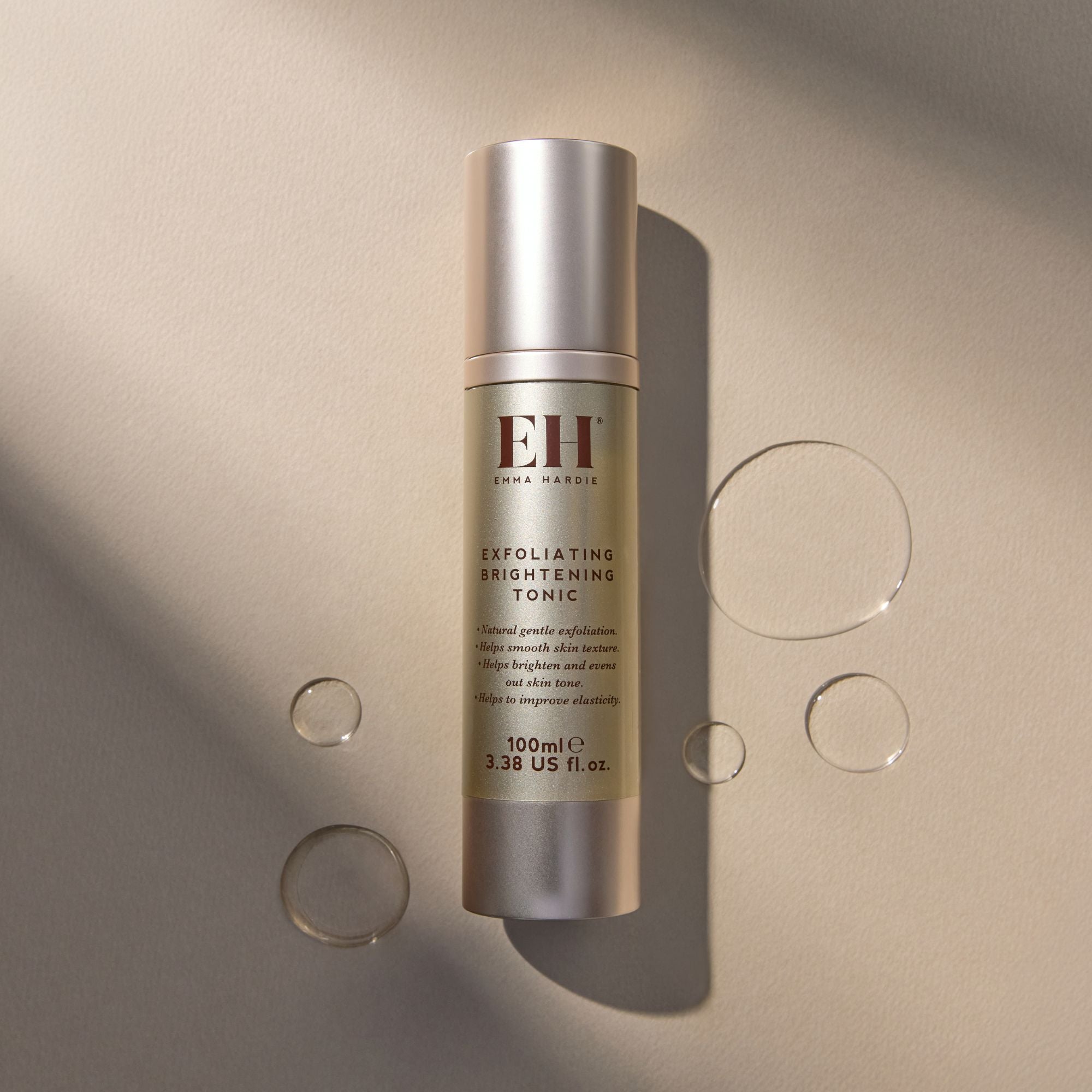 Emma Hardie Exfoliating Brightening Tonic bottle on a soft neutral background, surrounded by clear droplet-style accents and gentle shadows.