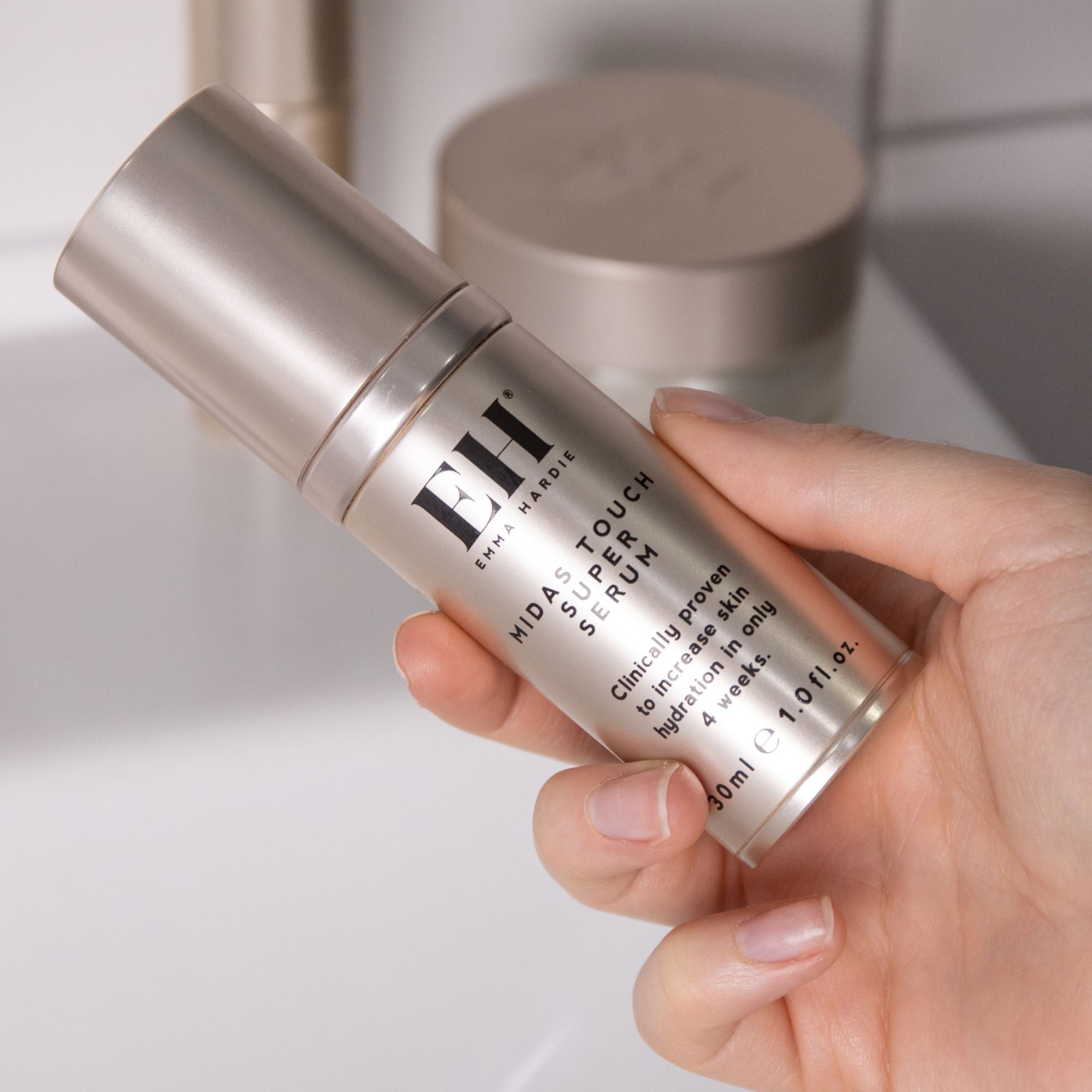 Hand holding Emma Hardie Midas Touch Super Serum bottle in a bathroom setting, ready for application.