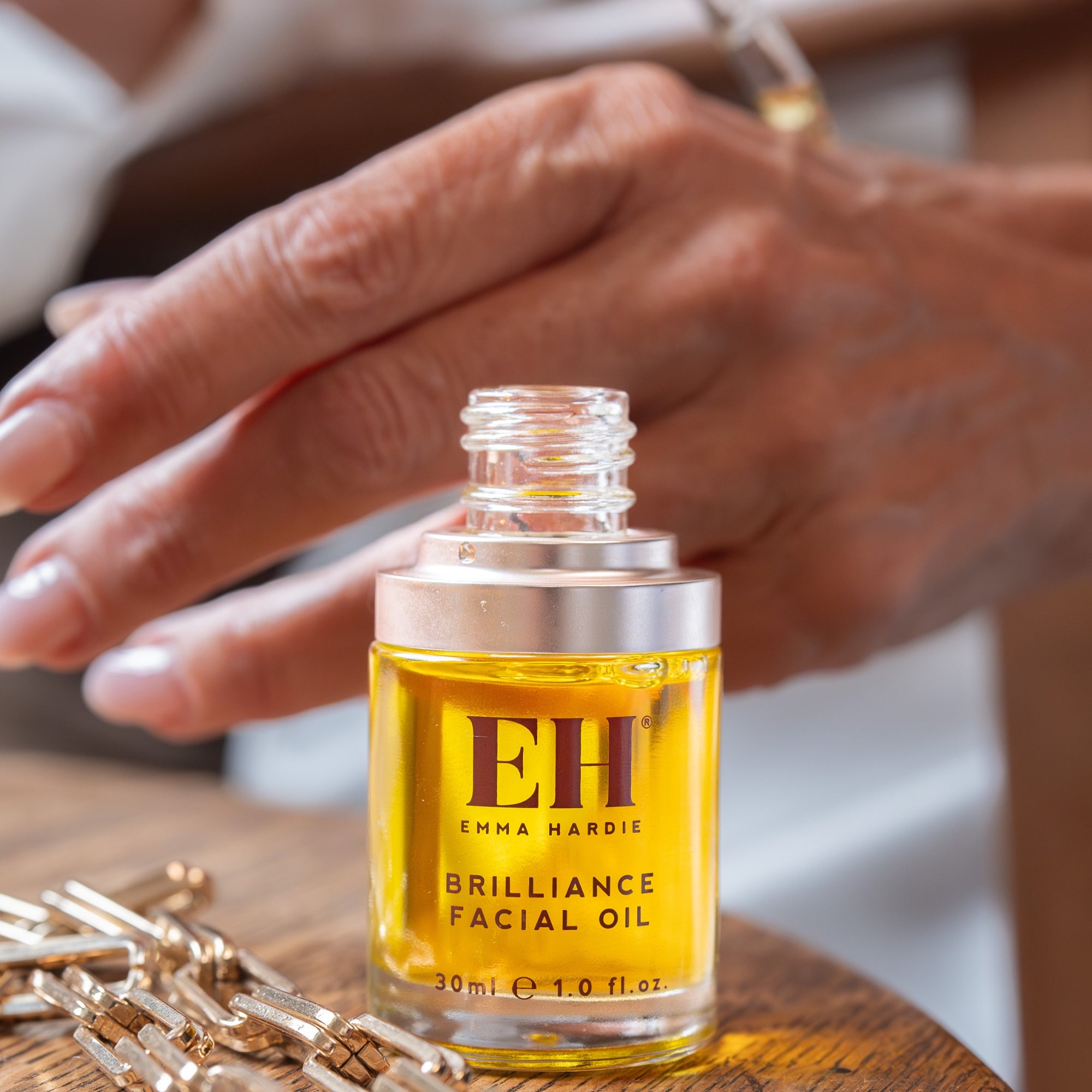 EMMA HARDIE Brilliance Facial Oil bottle on a wooden surface with hands in the background