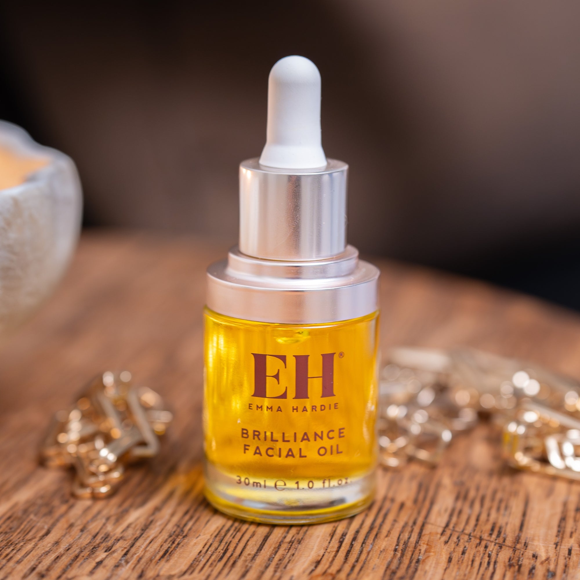 Bottle of Emma Hardie Brilliance Facial Oil on a wooden surface with jewelry in the background
