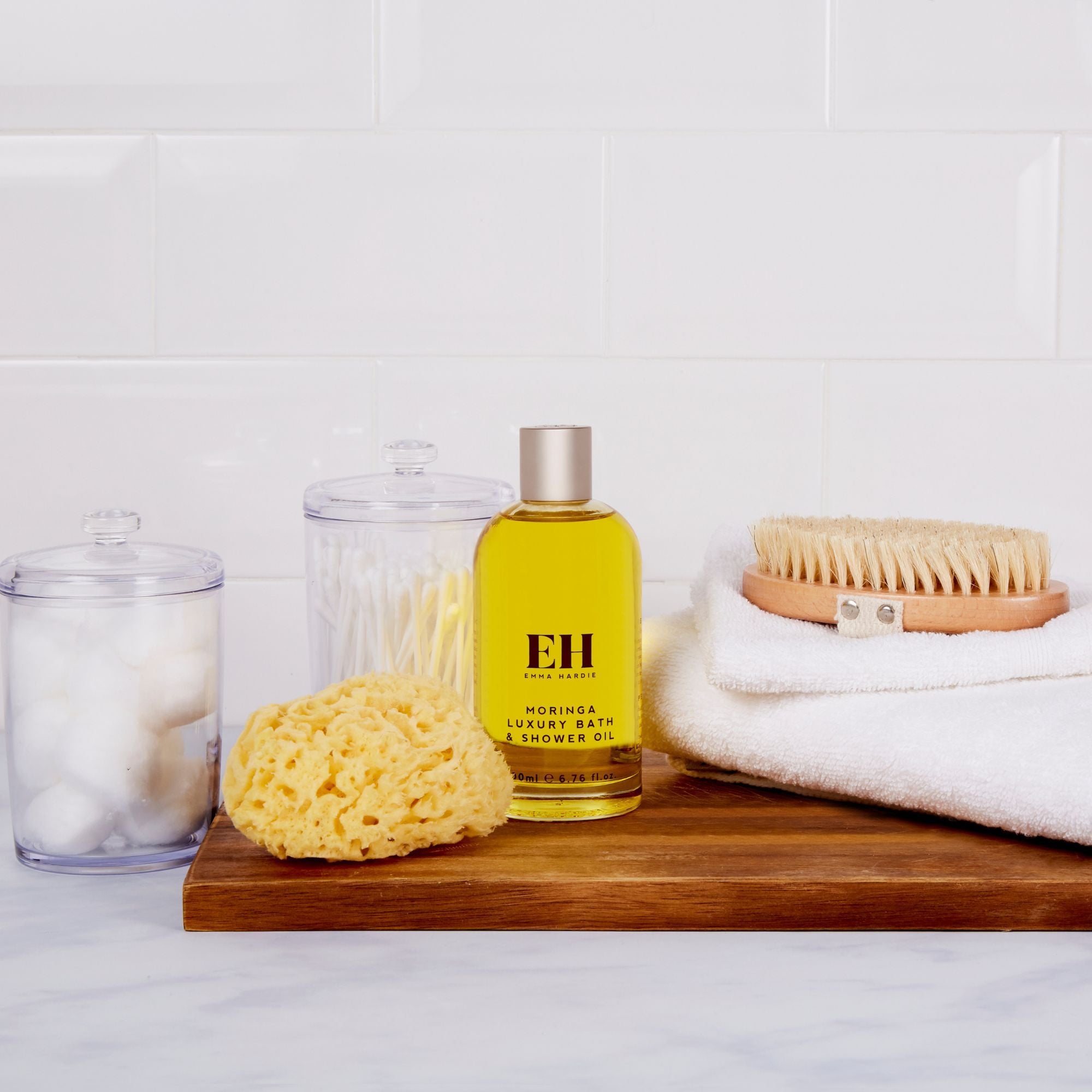 Bottle of EH Moringa Shower Oil on a wooden tray with a sponge, towel, and jars in the background.