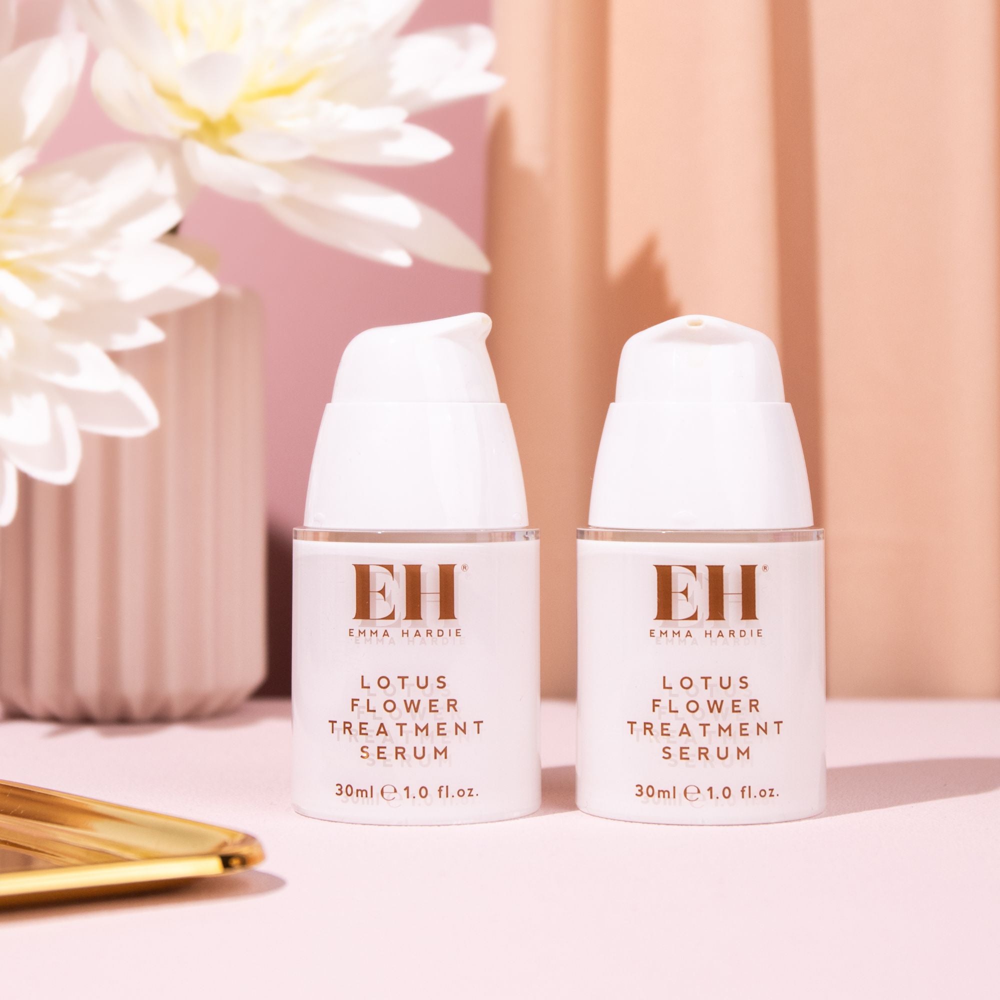 Two Emma Hardie Lotus Flower Treatment Serum bottles displayed on a soft pink background with white flowers and gold styling details.