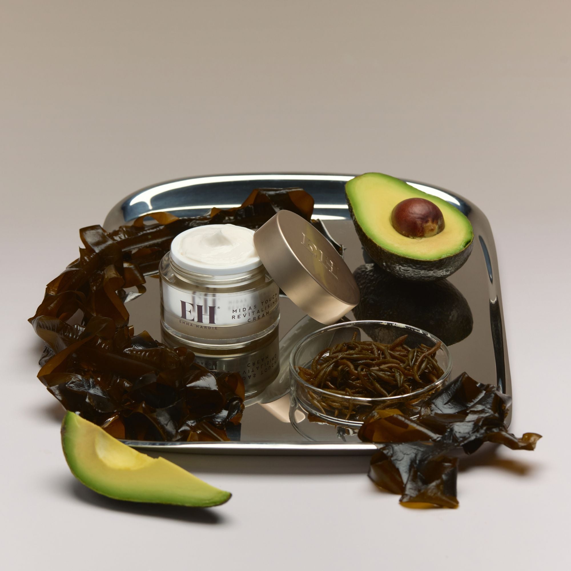 Silver tray with open jar of Emma Hardie Midas Touch Revitalising cream, avocado slices, and seaweed on a neutral background