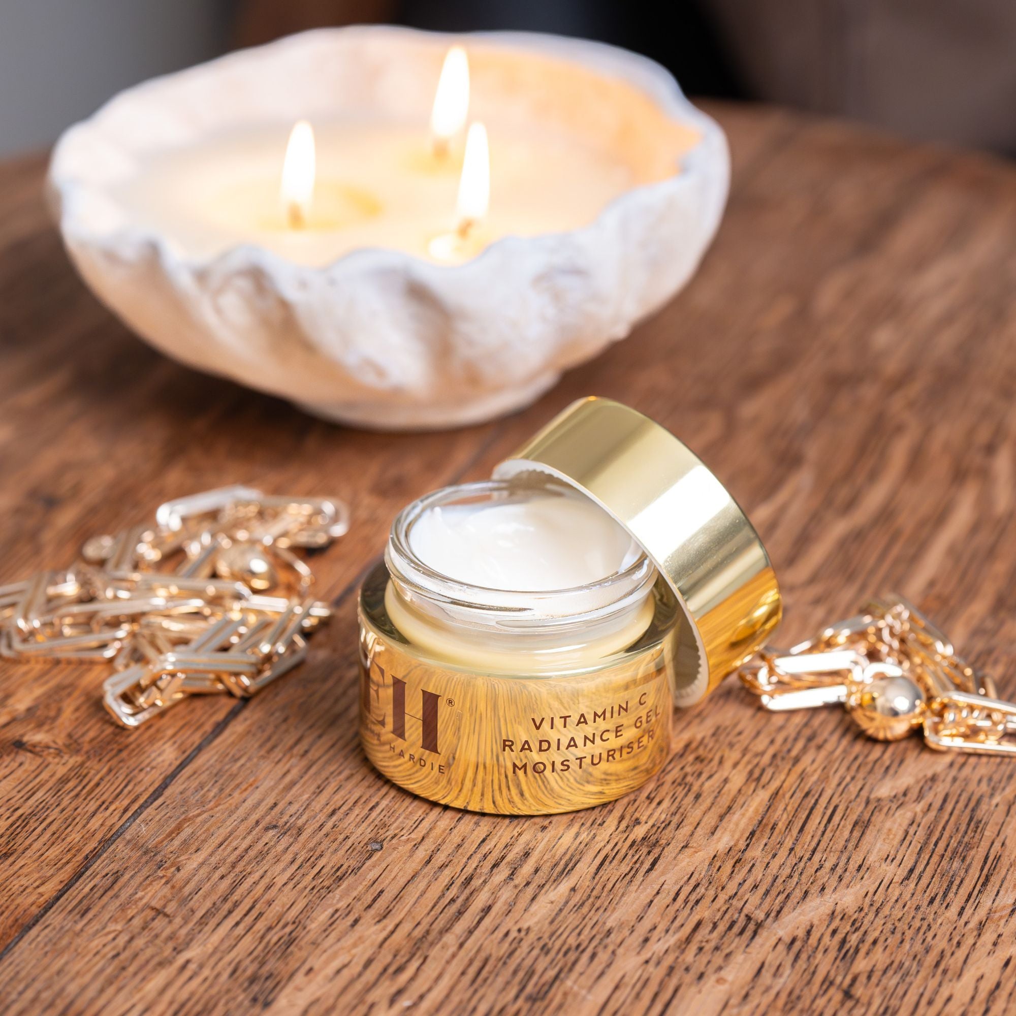 Open jar of Emma Hardie Vitamin C Radiance Gel Moisturiser on a wooden surface, styled with a glowing candle and gold chain details.