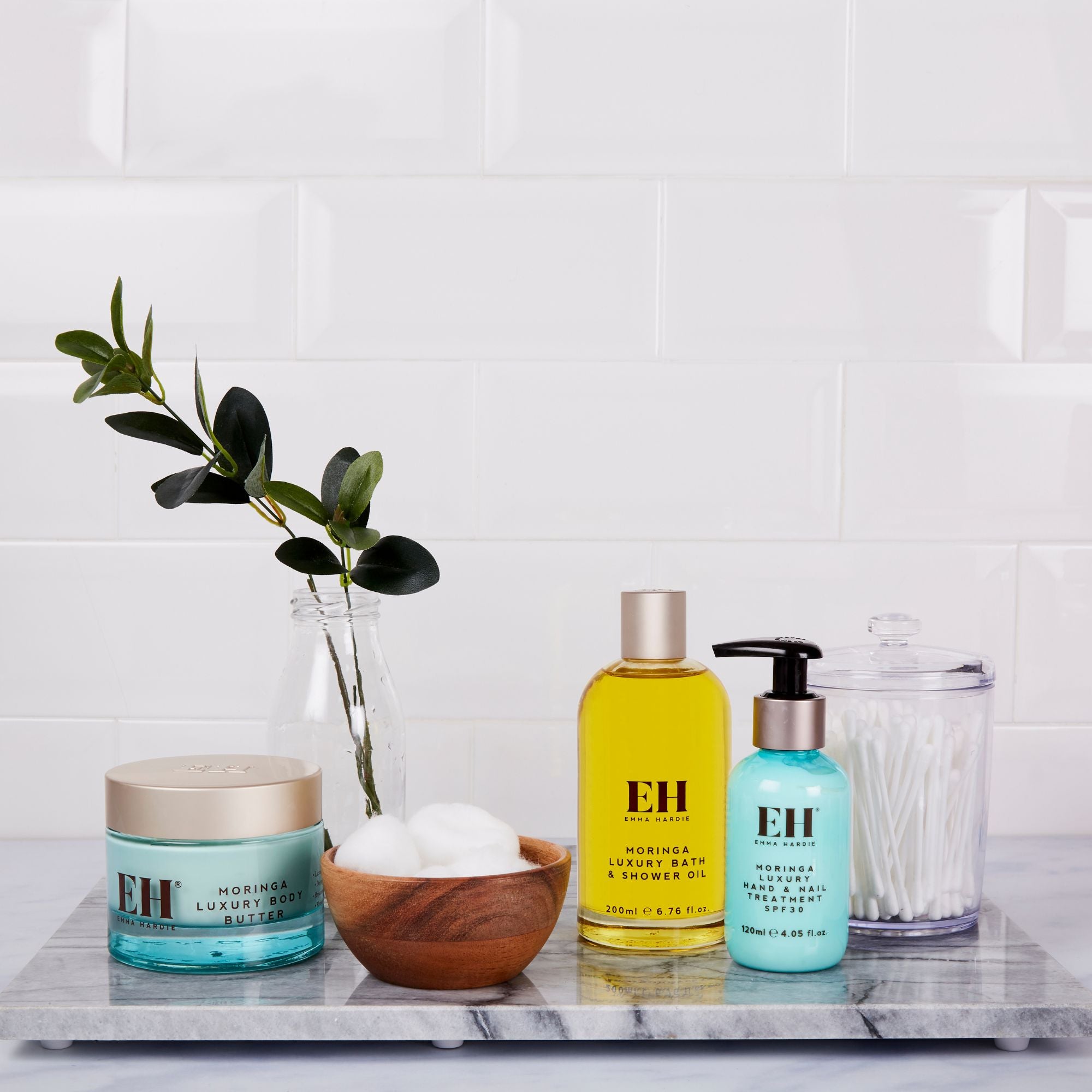 Emma Hardie Moringa body care products arranged on a marble tray with cotton pads, cotton buds and greenery in a bathroom setting.
