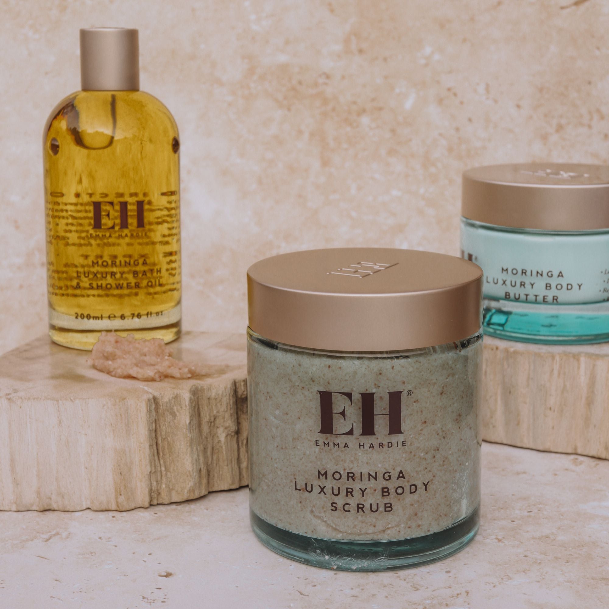 Emma Hardie Moringa Luxury Body Scrub displayed with Moringa Bath & Shower Oil and Body Butter on neutral stone plinths.