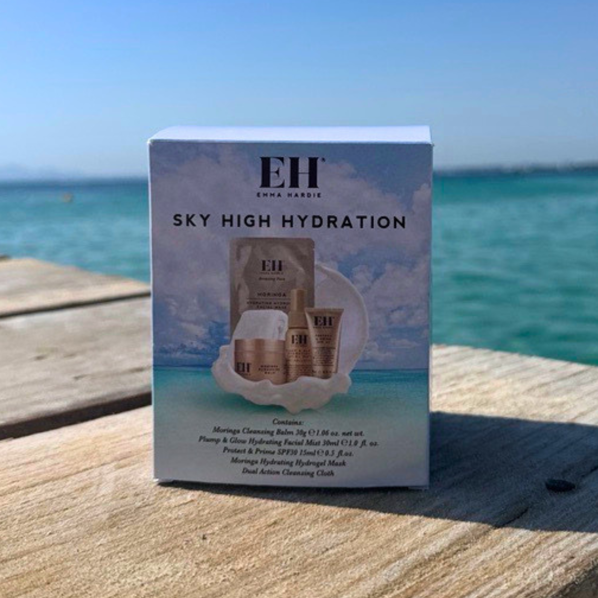 Emma Hardie Sky Hygh Hydration set on a wooden deck with turquoise water and blue sky in the background. The box shows the EH logo and an illustration of the included items, including a facial mask, toner, and cream bottles.