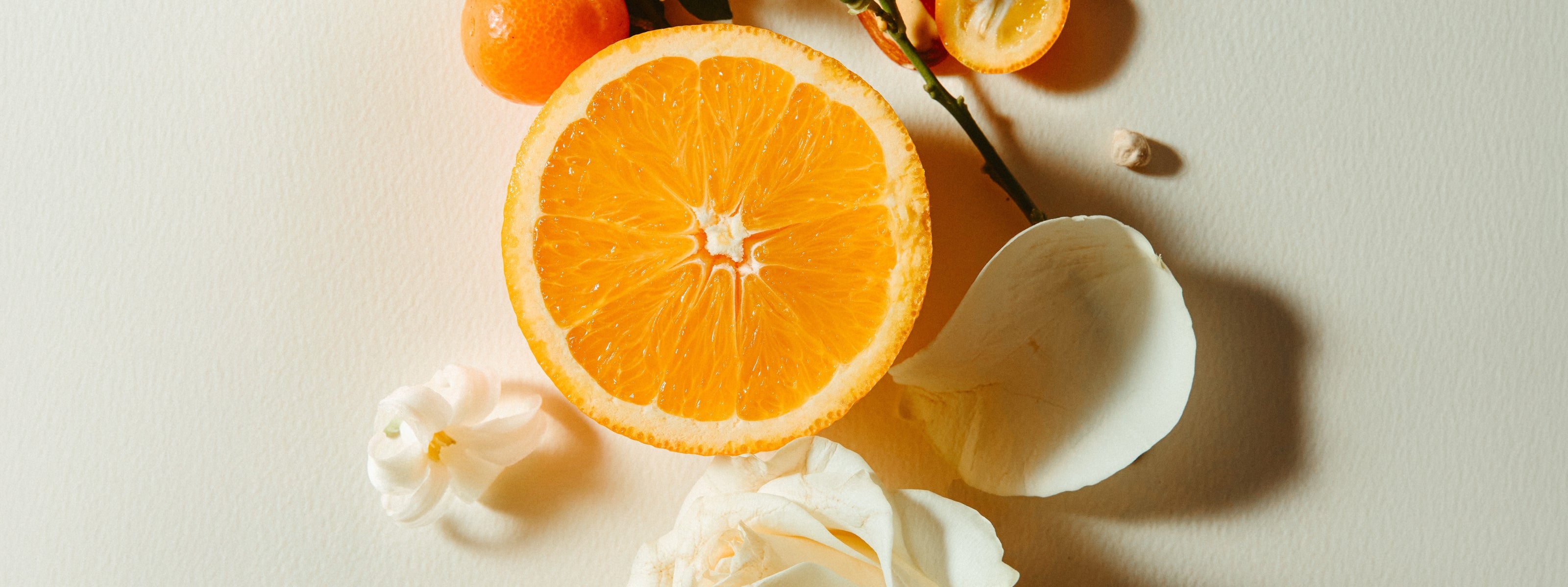 A styled flat-lay of a halved orange, small orange fruits on a sprig with green leaves, almonds, and white flower petals arranged around a large white rose on a warm beige background.