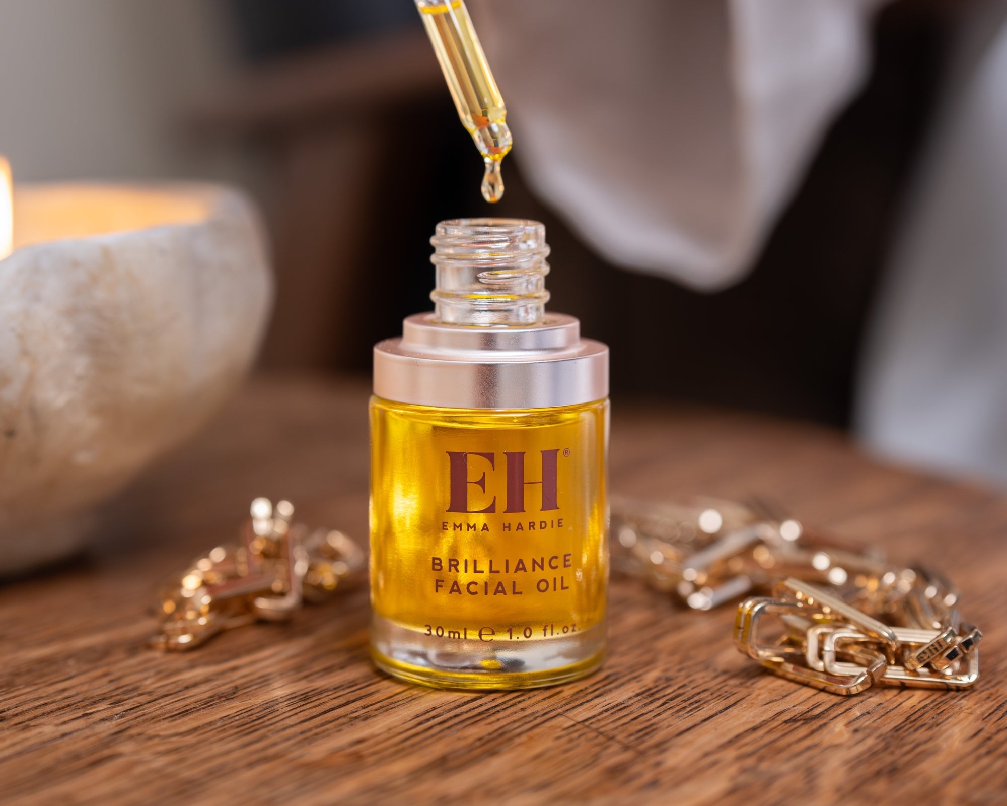 Emma Hardie Brilliance Facial Oil with dropper above the open bottle, displayed on a wooden table with gold accessories.