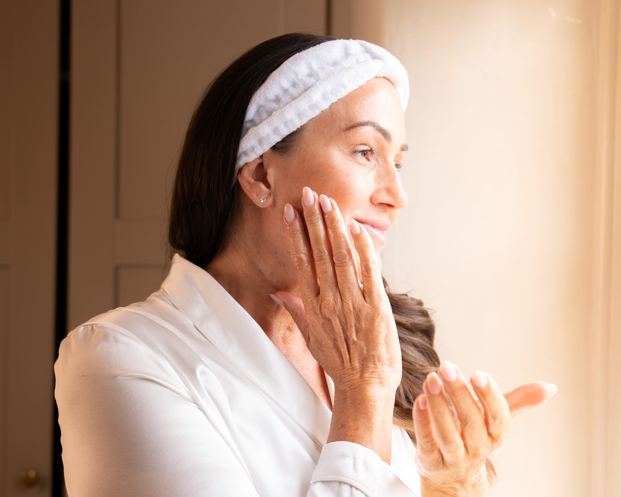 Woman wearing a skincare headband gently massaging product into her cheek as part of an at-home facial ritual.