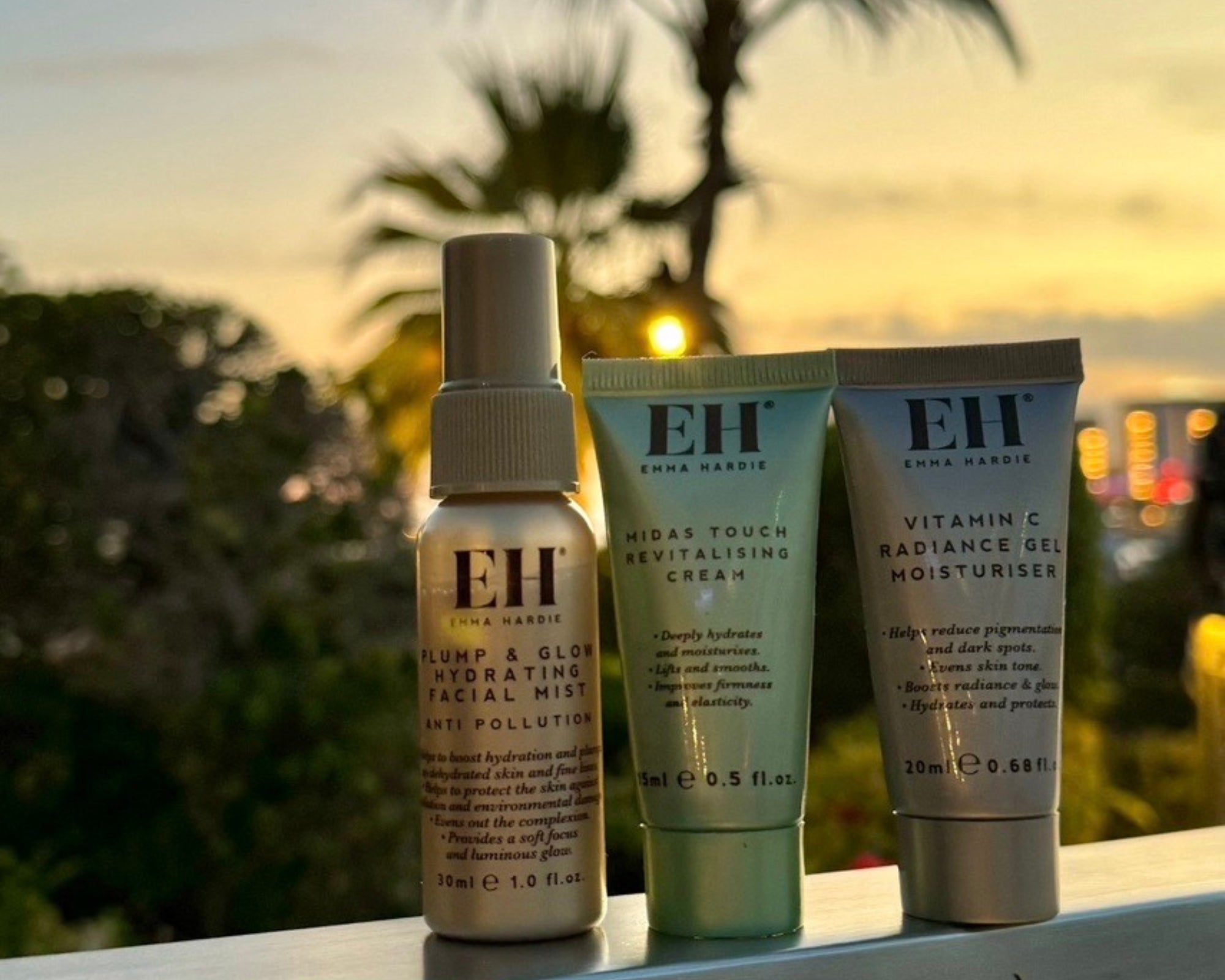 Emma Hardie travel-size Plump & Glow Hydrating Facial Mist, Midas Touch Revitalising Cream and Vitamin C Radiance Gel Moisturiser against a sunset backdrop.
