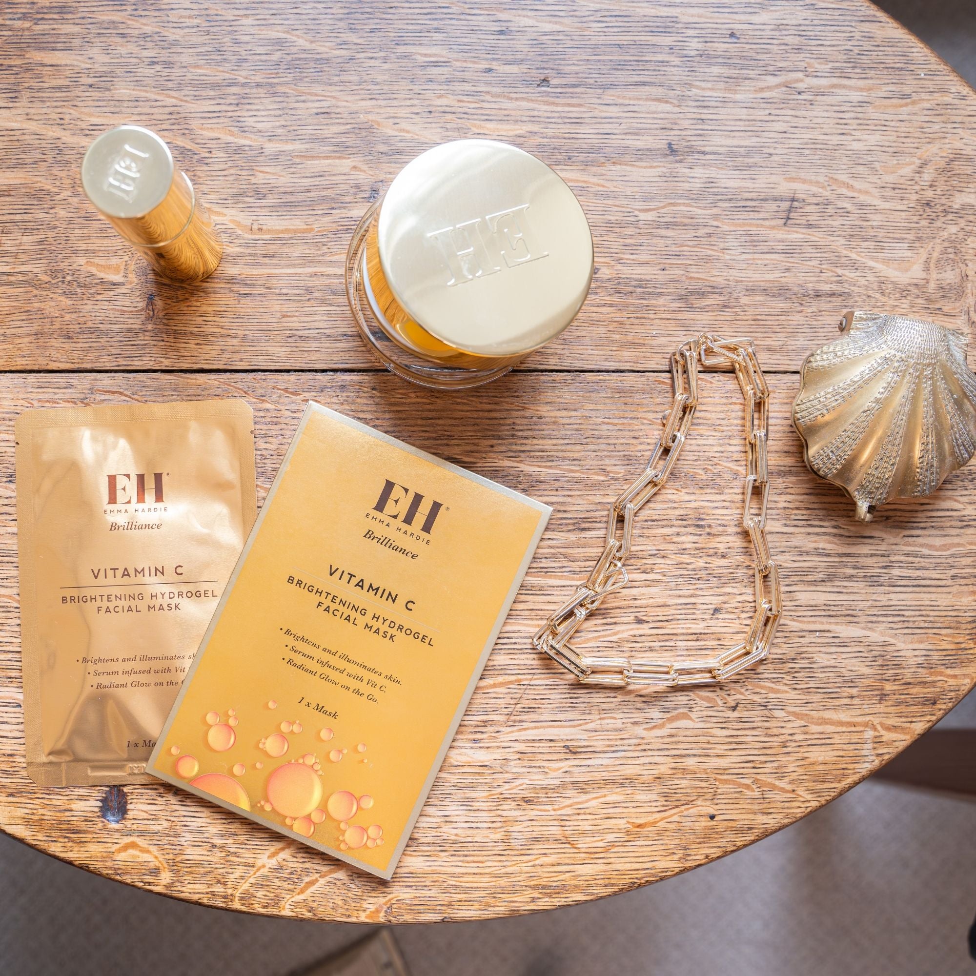 Emma Hardie Vit C Brilliance Cleansing Balm, Radiant Renewal Serum and Brightening Hydrogel Facial Mask on a wooden table.