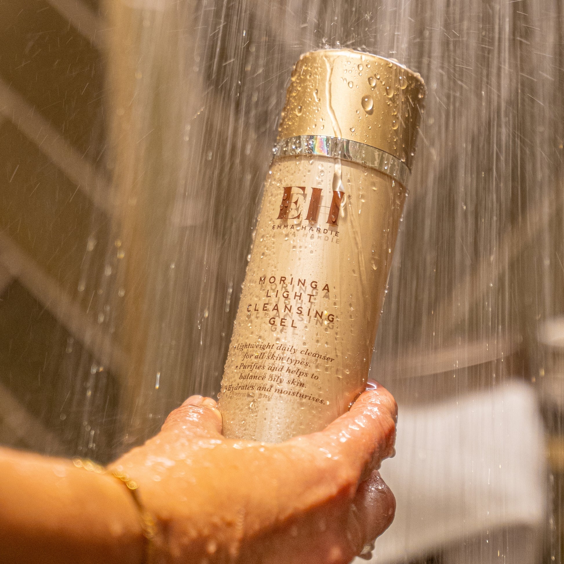 Hand holding Emma Hardie Moringa Light Cleansing Gel under running shower water, with water droplets across the bottle.