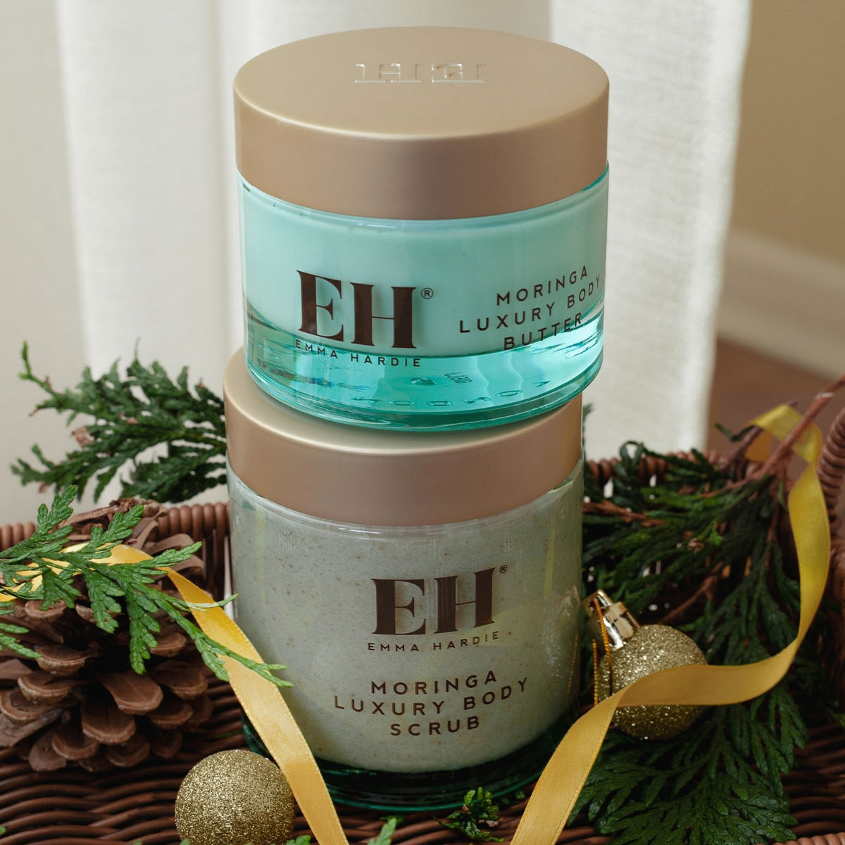 Emma Hardie Moringa Luxury Body Butter stacked on Moringa Luxury Body Scrub in a wicker basket with festive greenery, gold baubles and ribbon.
