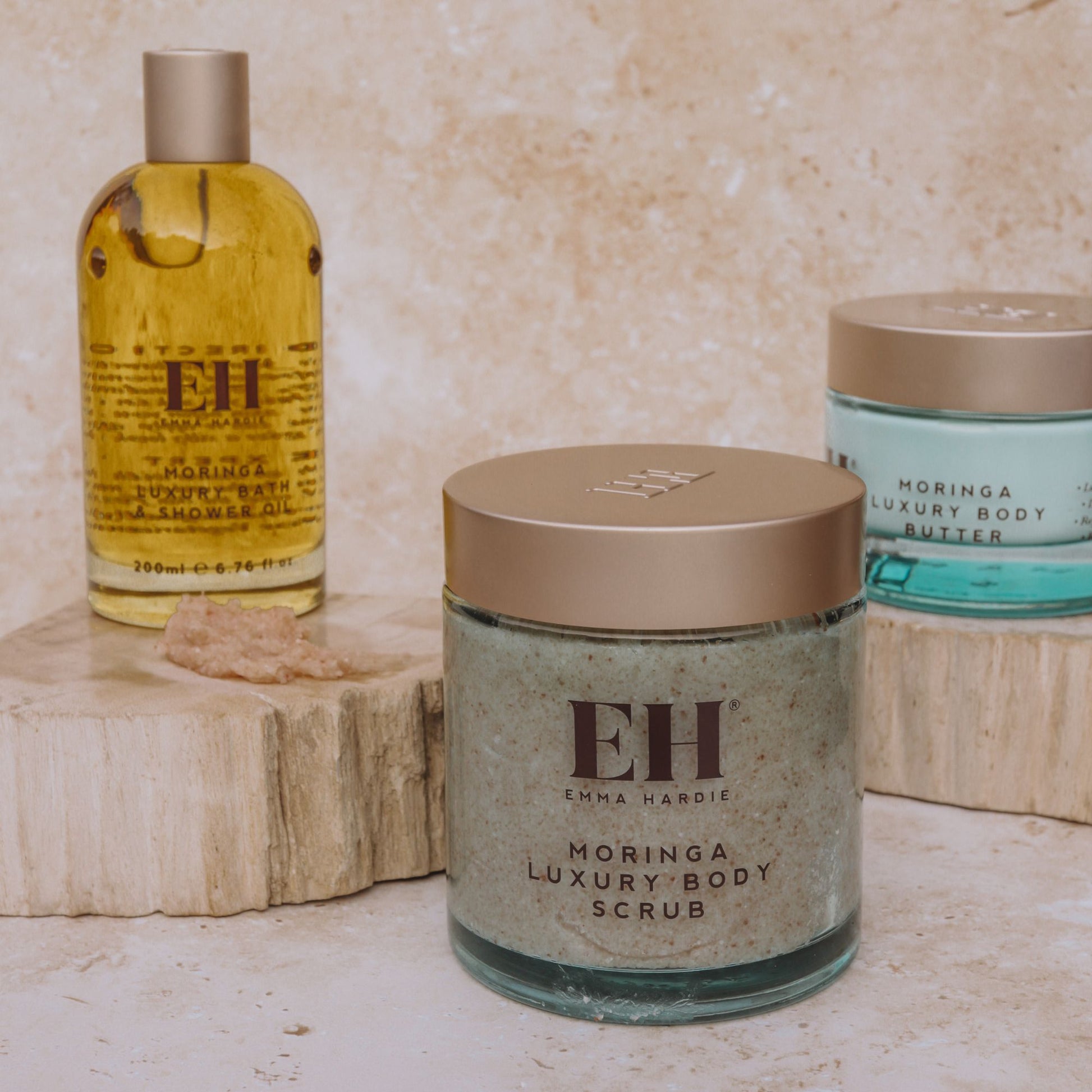 Emma Hardie Moringa Luxury Body Scrub displayed with Moringa Bath & Shower Oil and Body Butter on neutral stone plinths.